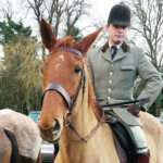 A young man in tweed hunting attire mounted on a light-brown horse, with riders and cars in the background.