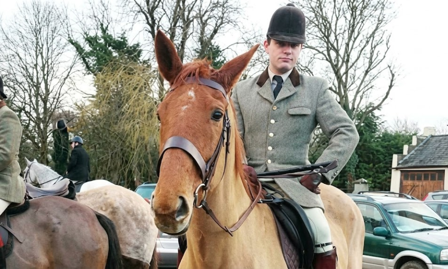 A young man in tweed hunting attire mounted on a light-brown horse, with riders and cars in the background.
